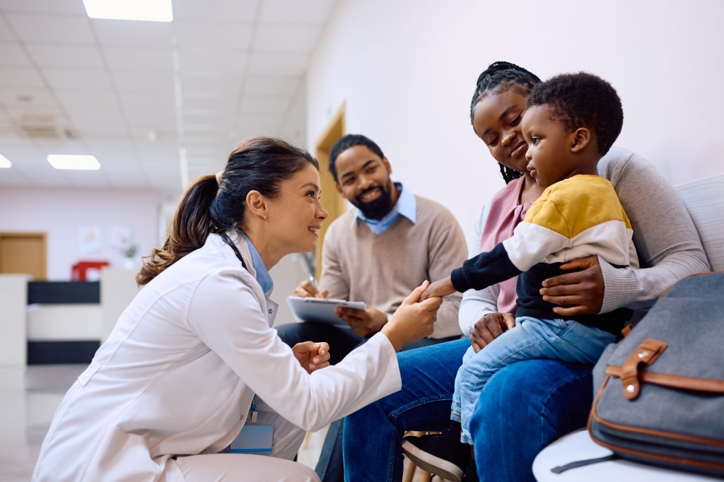 Female pediatrician talking to black little boy who is sitting on mother's lap at the clinic.