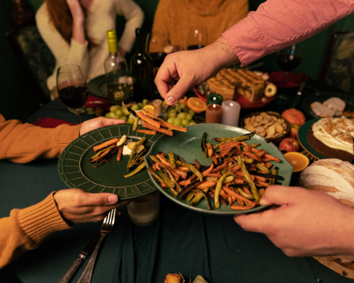 Unidentified people's arms and hands shown serving roasted vegetables on a green plate to other unidentifiable people seated at the table. The table has various a variety of traditional Thanksgiving dishes such as bread, vegetables, and pie. 