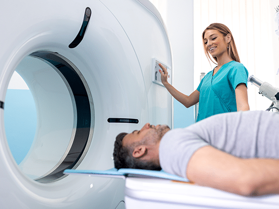 Woman in teal scrubs is smiling at a patient as she operates a large medical imaging machine. The patient is laying on their back inside the machine. 