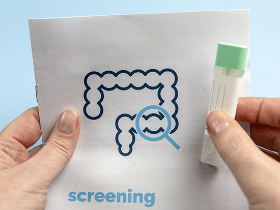 A person holds an at-home colon cancer screening kit, featuring a test tube with a green cap and an instructional pamphlet displaying a stylized image of a colon with a magnifying glass.