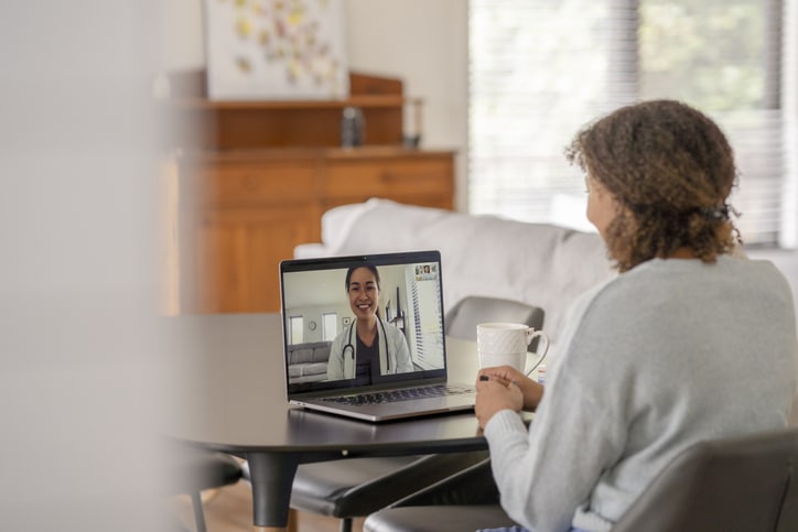 A female patent meets with her doctor remotely via a video call. She is sitting at her kitchen table with her laptop out and a cup of coffee on the table with her. The doctor can be seen on the screen wearing a white lab coat as she talks with her patient.
