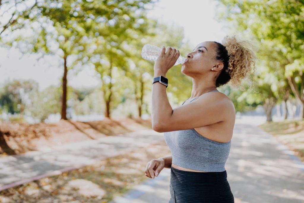 Woman wearing athletic clothing drinks from a water bottle while walking outdoors on a sunny, tree-lined path, illustrating the importance of staying hydrated during summer exercise.