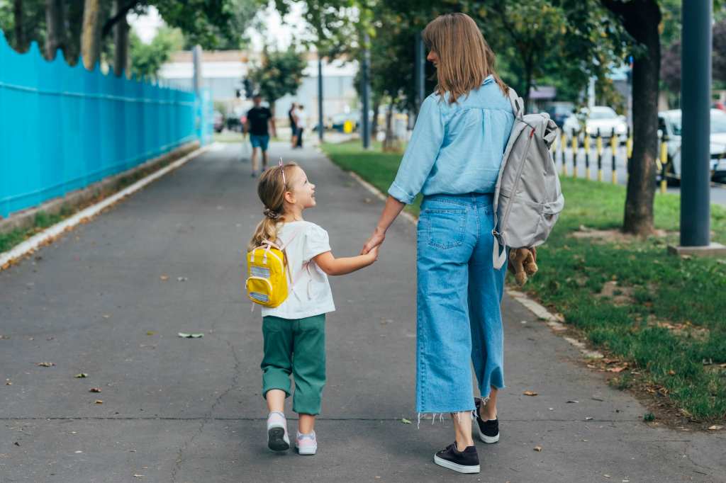 Mother walking her young daughter with a yellow backpack to school, holding hands on a sidewalk, symbolizing back-to-school season and family health.