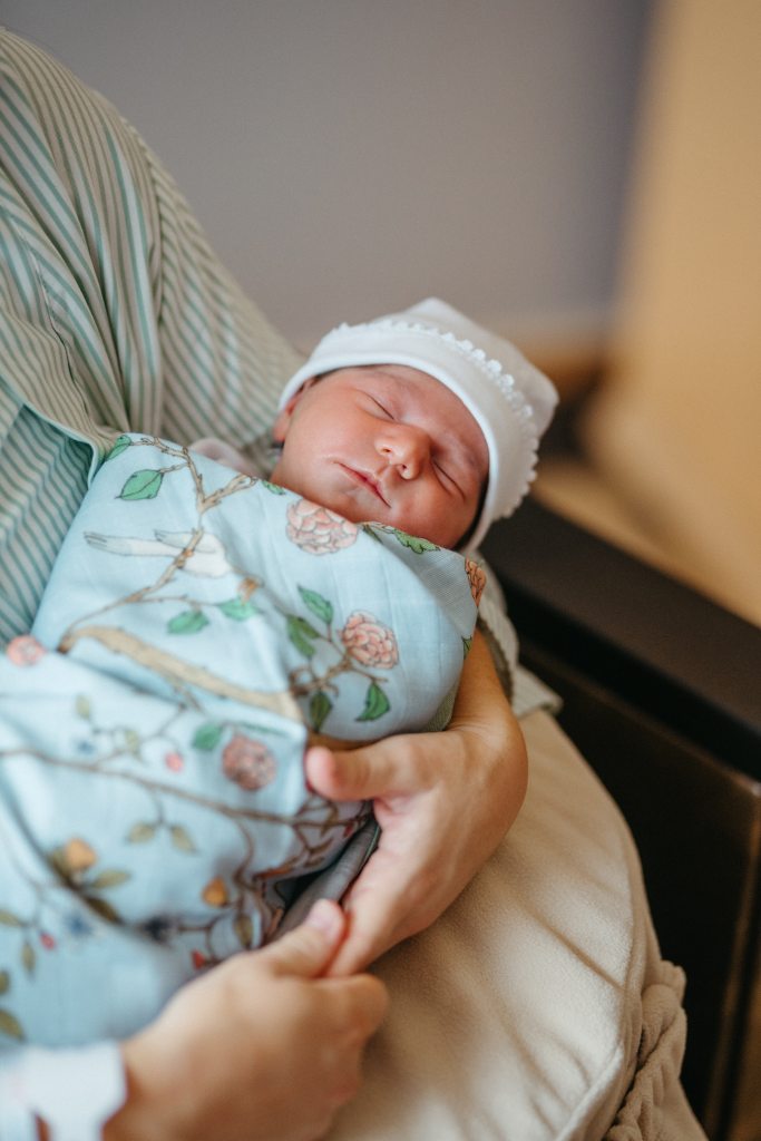A newborn baby sleeps safely on its back in a caregiver’s arms, wrapped in a floral blanket and wearing a white cap, demonstrating safe sleep practices recommended by the American Academy of Pediatrics.