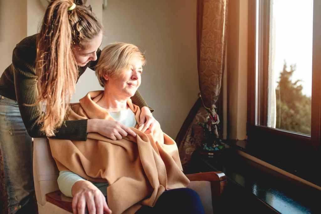 A caregiver gently wraps a blanket around an older adult sitting by a sunny window, symbolizing compassionate senior care and caregiver support at home.
