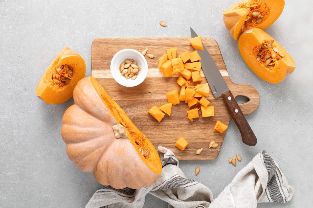 Fresh pumpkin cut into slices and cubes on a wooden cutting board with a knife and bowl of seeds, illustrating healthy cooking and nutrition benefits of pumpkin.