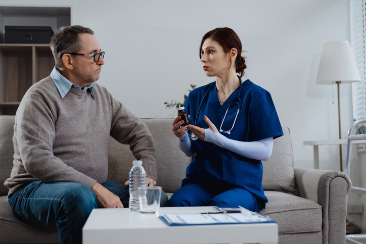 A nurse in blue scrubs sits on a couch and talks with a middle-aged man, showing him a medical device.