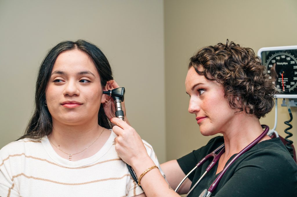A primary care clinician uses an otoscope to examine a patient’s ear during a routine checkup in a UAB St. Vincent’s exam room. The patient sits calmly while the clinician performs the ear exam, with medical equipment visible in the background.