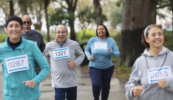 A group of five adults running in a race in a park. They are wearing numbered sports bibs. The focus is on the man in the middle, a senior in his 60s. He is looking at the camera.