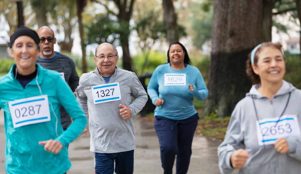 A group of five adults running in a race in a park. They are wearing numbered sports bibs. The focus is on the man in the middle, a senior in his 60s. He is looking at the camera.