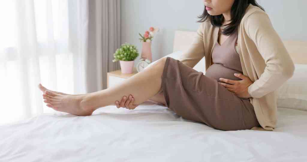 Pregnant woman sitting on a bed and holding her lower leg while experiencing discomfort, highlighting common symptoms of varicose veins during pregnancy.