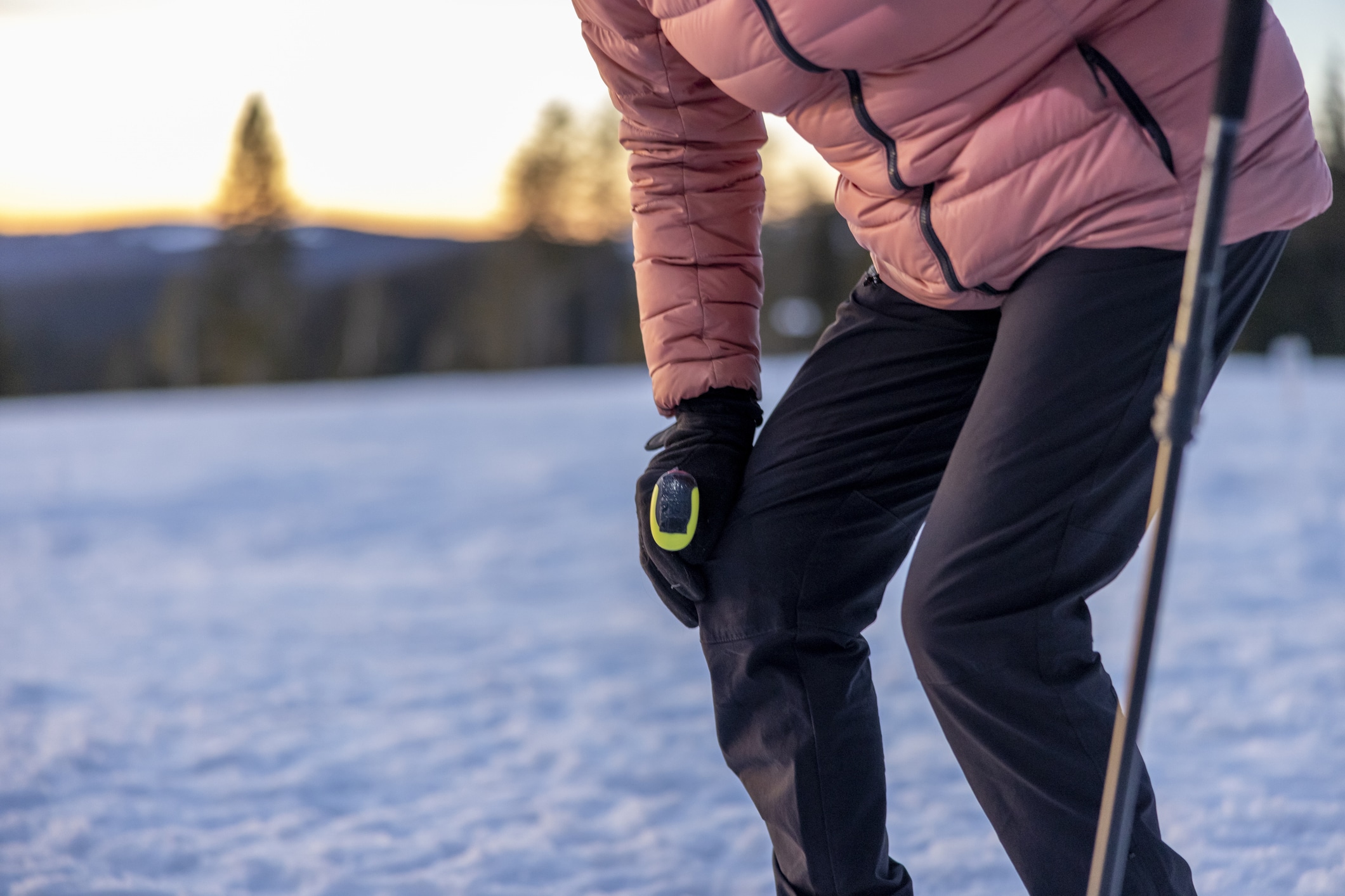 An individual outdoors in cold weather pauses to hold their knee, showing signs of joint discomfort while standing on a snowy landscape at sunset.
