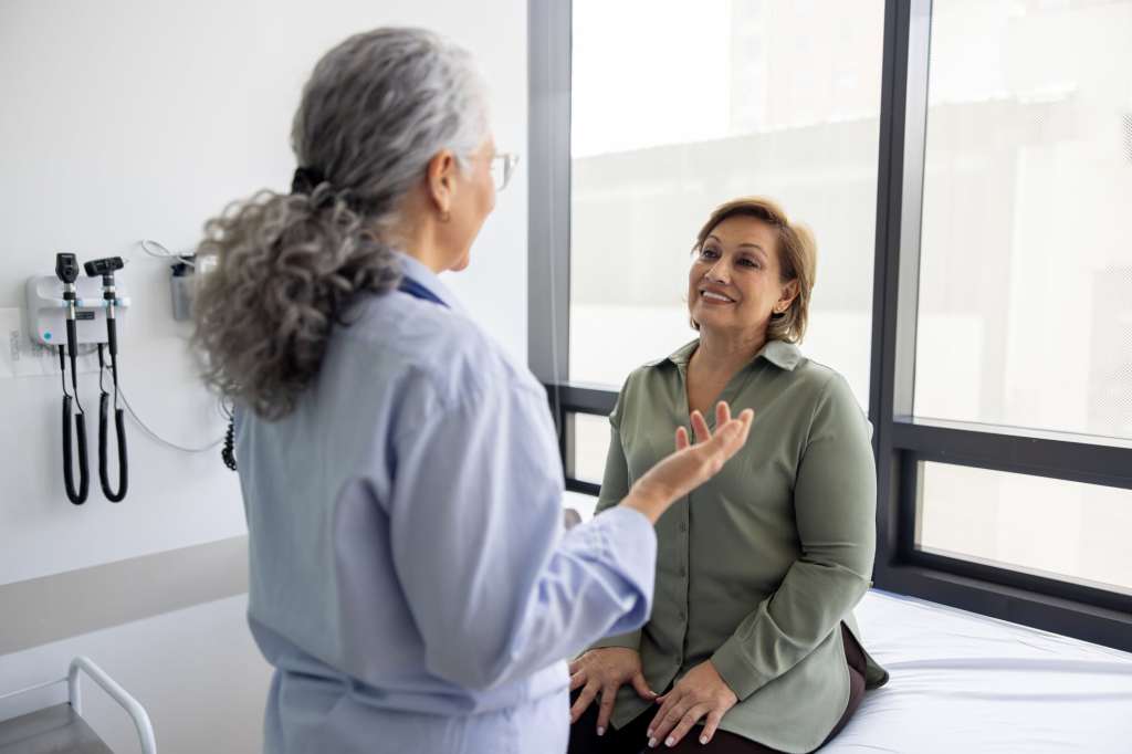 Health care provider speaking with a middle-aged woman during a medical visit, discussing perimenopause symptoms and women’s health care in a clinical exam room.