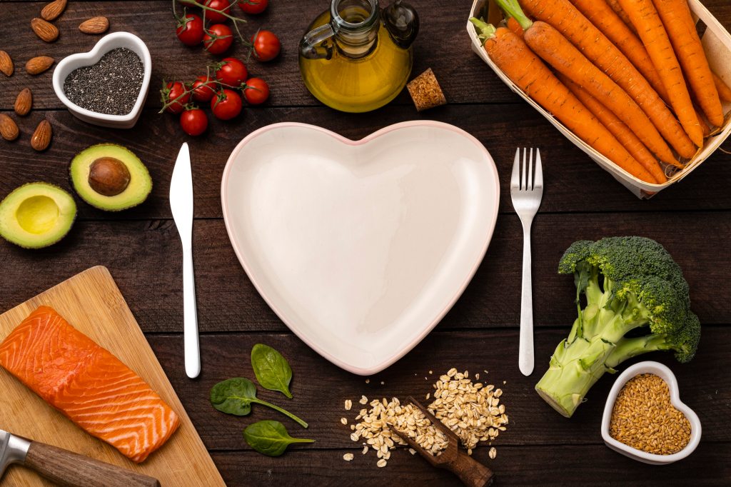 Top view of a heart shaped plate surrounded by a knife and a fork and some healthy food such as a salmon fillet, broccoli, oat flakes, chia and flax seeds, cherry tomatoes, carrots, almond, avocado, spinach and olive oil. All the objects are on a rustic wooden table.