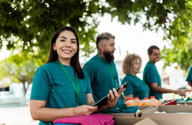 Volunteers standing outdoors under trees distribute supplies from boxes at a community event while a woman in a green shirt holds a tablet and smiles at the camera.