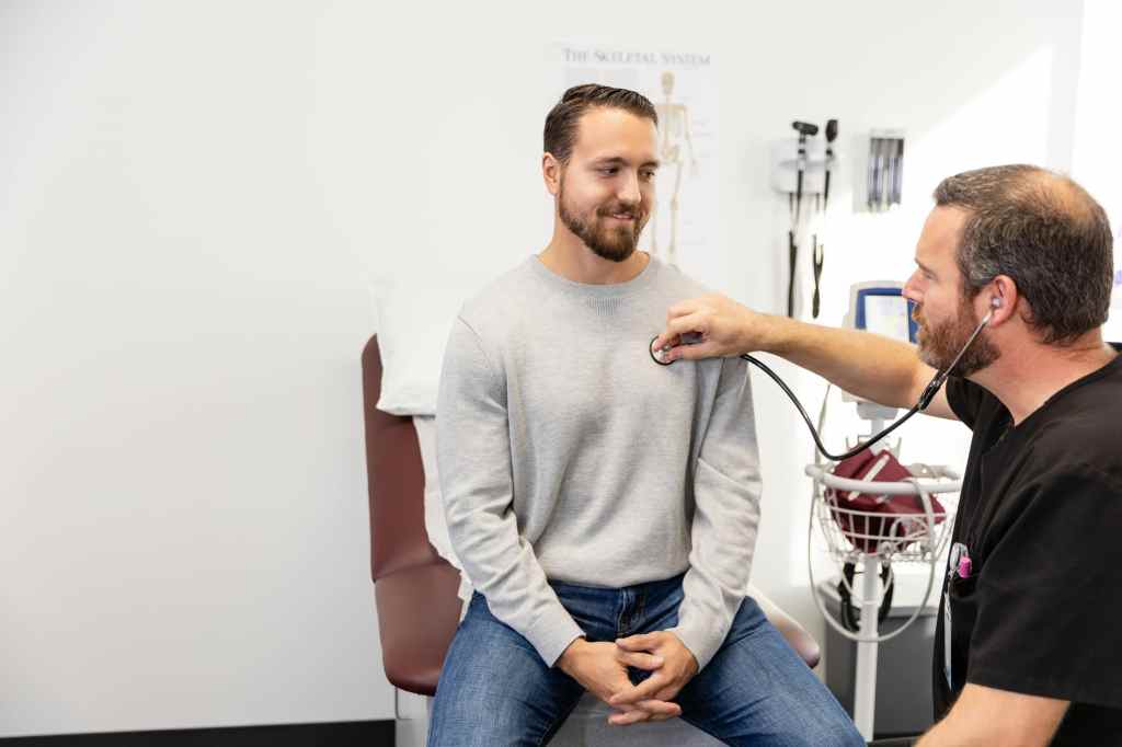 Male patient sits while doctor uses a stethoscope during a medical checkup in a clinic.