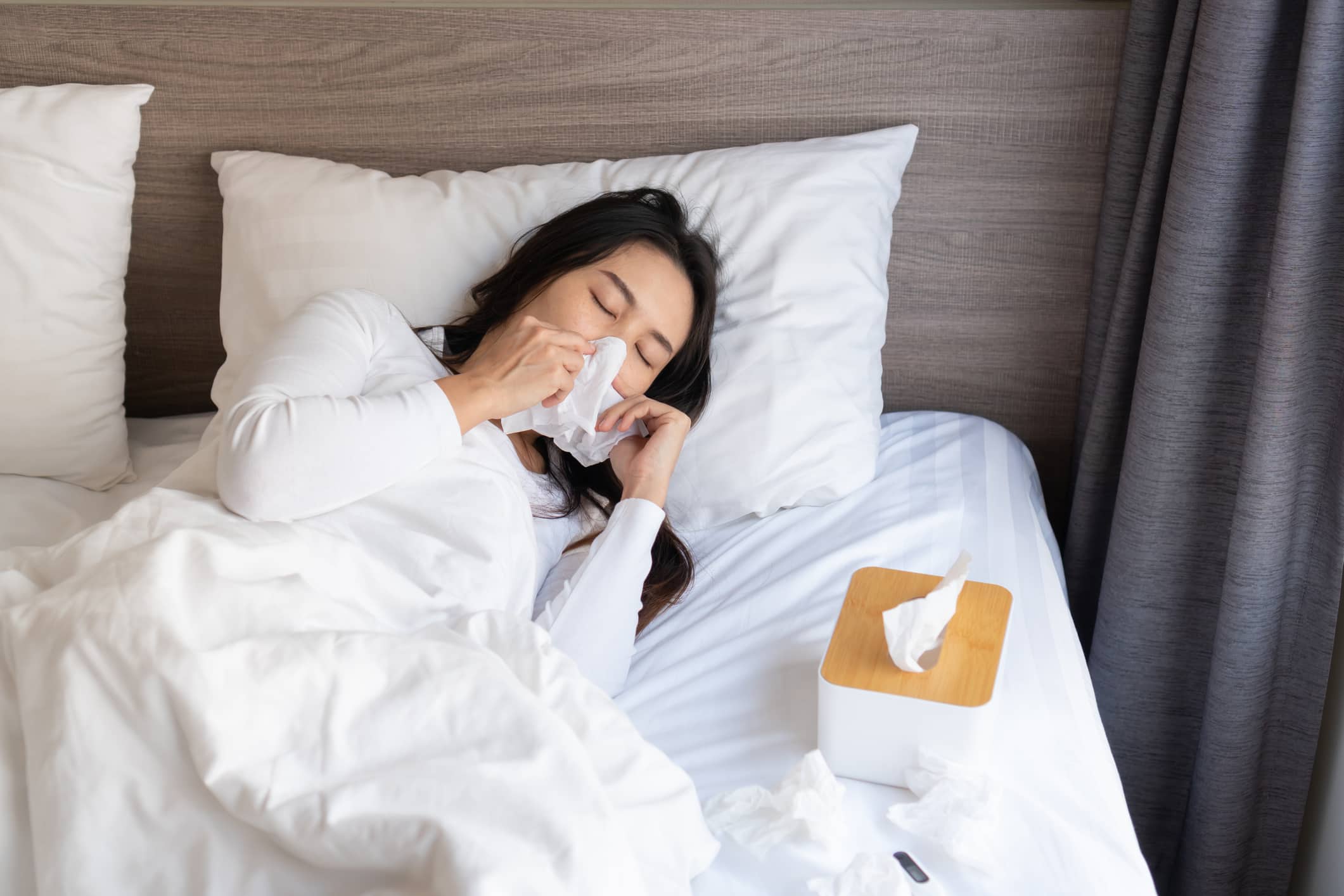 Young woman sneezing from allergy symptoms blowing nose on bed after wakeingup in the morning. 