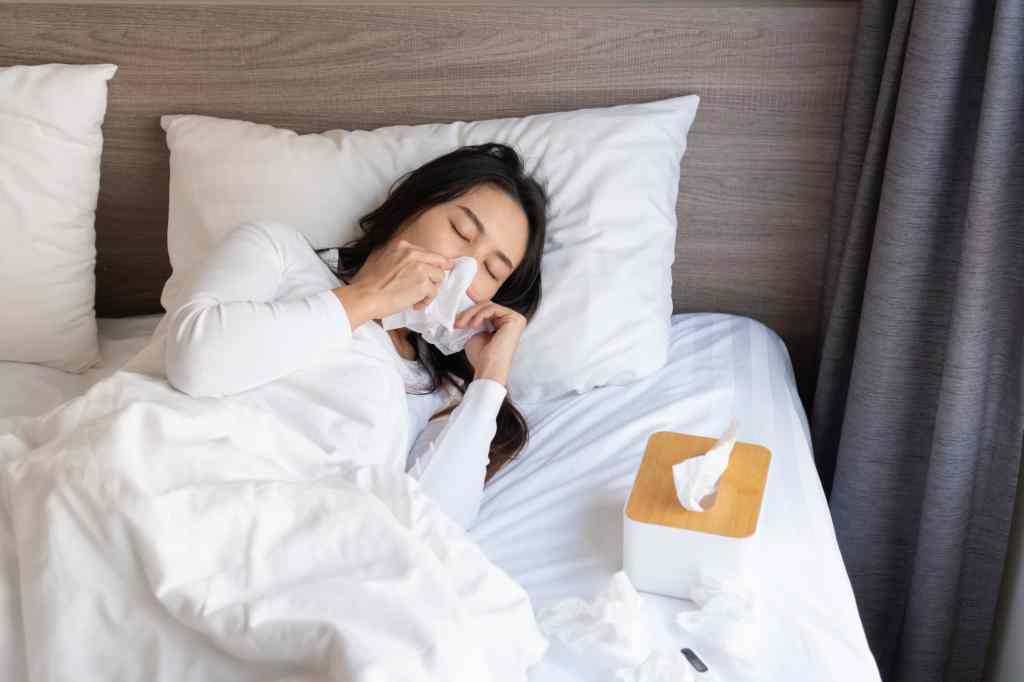 Young woman sneezing from allergy symptoms blowing nose on bed after wakeingup in the morning.