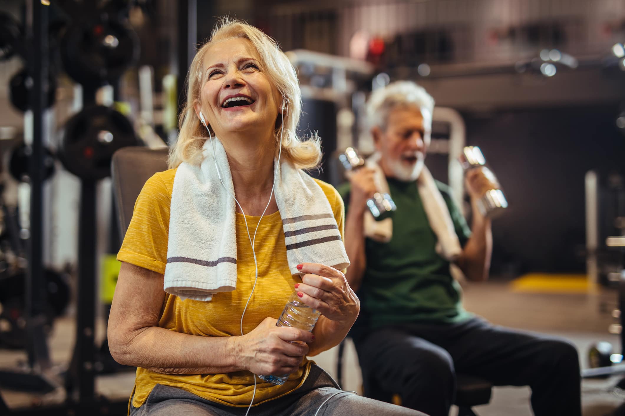Woman sitting at weight machine in a gym. She has headphones in while smiling with a sweat towel around her neck