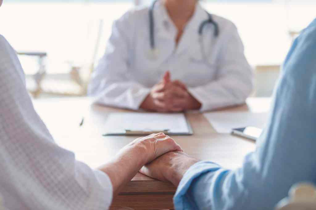 couple holding hands at a doctors office. Doctor can be seen in the background.